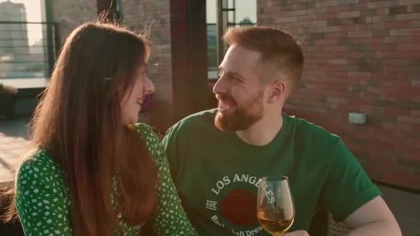 Giovane coppia uomo e donna con bicchieri di vino in mano a chiacchierare e mangiare sushi rotoli seduti sulla terrazza con vista sulla città - Filmati, video