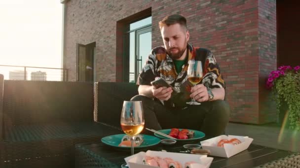 Jonge aantrekkelijke man met een glas wijn in de hand het eten van sushi rollen zittend op terrasvormige dek - Video