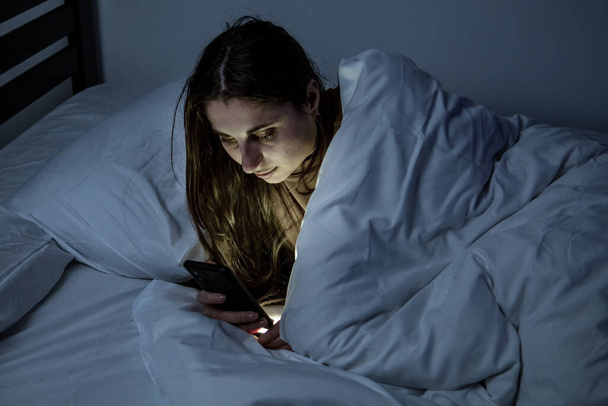 Dívka v posteli s telefonem na bílé posteli v tmavé místnosti - Fotografie, Obrázek