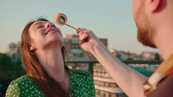 Close-up of a guy holding a delicious fresh roll of sushi with chopsticks on terrace feeding his girlfriend - Footage, Video