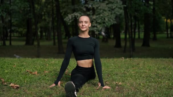 Een jong mooi meisje zit op de splits en doet stretching na de training in de ochtend in het park - Video