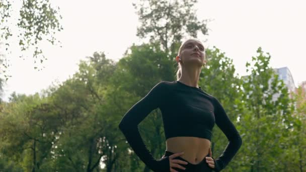 девушка в спортивной одежде делает круговые движения шеей во время разминки перед тренировкой в парке - Кадры, видео