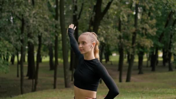 portret van een jong mooi meisje in sportkleding in een stadspark doen stretching oefeningen voor de spieren de armen sport training - Video