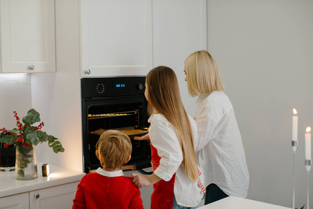 Äiti ja lapset leipovat joulupiparkakkuja modernissa keittiössä. Äiti laittaa keksejä sisältävän tarjottimen uuniin. Perheloman valmistelu - Valokuva, kuva