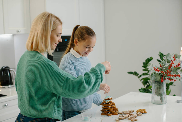 Mère heureuse et fille mignonne fille décorant biscuits de pain d'épice de Noël après la cuisson tout en se tenant dans la cuisine confortable à la maison avec des décorations de Noël. - Photo, image