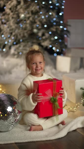 A lány egy piros dobozt tart egy ajándékkal a karácsonyfa közelében, az újévi fények hátterében. Egy boldog gyermek, aki ajándékot kapott.. - Felvétel, videó