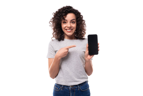 junge lustige hübsche brünette lockige kaukasische Dame in lässigen Jeans und T-Shirt demonstriert einen Smartphone-Bildschirm mit einer Attrappe für Werbung auf weißem Hintergrund mit Kopierraum. - Foto, Bild