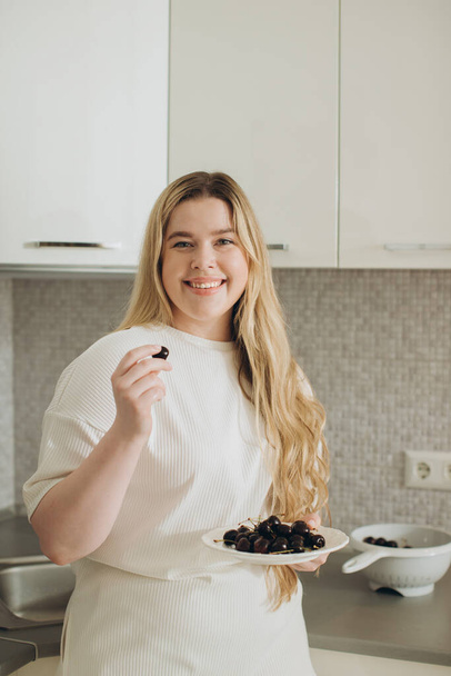 Nuori hymyilevä nainen syö tuoreita kirsikoita keittiössä - Valokuva, kuva