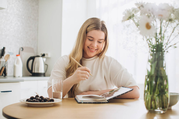 Portrait d'une jolie étudiante qui tient son journal tout en étant assise à la table dans la cuisine. - Photo, image