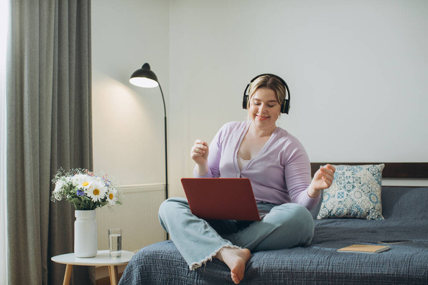 Девушка с светлыми волосами в наушниках смотрит развлекательное видео или слушает музыку на ноутбуке, сидя в постели в спальне дома. - Фото, изображение