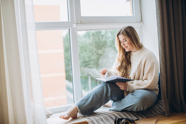 クリエイティブな若い女性は,自宅の窓の近くに座っている間,ノートブックにメモリプランとアイデアを書いています. クリエイティブジャーナルや日記コンセプト. - 写真・画像