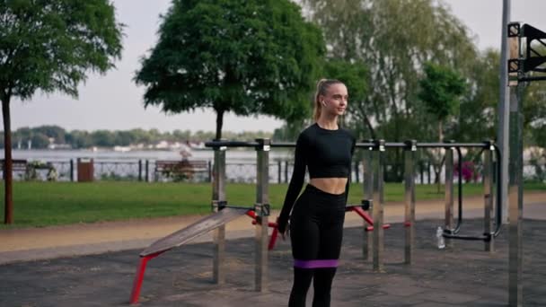 Όμορφη κοπέλα με αθλητική στολή εκτελεί ασκήσεις για να ανεβάσει τα πόδια της με λαστιχάκι στην παιδική χαρά του δρόμου σπορ - Πλάνα, βίντεο