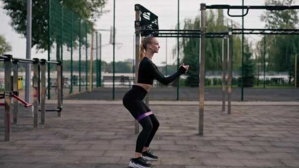 Belle fille en uniforme de sport effectue des exercices pour pomper ses jambes avec élastique sur l'aire de jeux de rue sportive - Séquence, vidéo