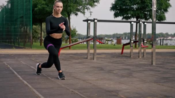 Menina bonita em uniforme esportivo realiza exercícios para bombear as pernas com elástico no playground de rua - Filmagem, Vídeo