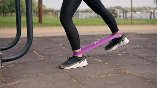 Primer plano de piernas de niña entrenadora deportiva realizando ejercicios con una banda elástica para fortalecer los músculos en el campo deportivo - Imágenes, Vídeo