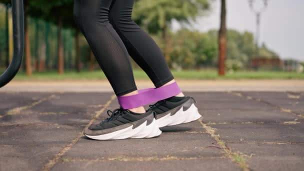 Κοντινό πλάνο των ποδιών του αθλητικού γυμναστή κορίτσι εκτελεί ασκήσεις με μια ελαστική ζώνη για την ενίσχυση των μυών στο πεδίο του αθλητισμού - Πλάνα, βίντεο