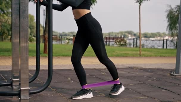 Κοντινό πλάνο των ποδιών ενός κοριτσιού αθλητικό εκπαιδευτή εκτελεί ασκήσεις με ένα ελαστικό ιμάντα για την ενίσχυση των μυών στο πεδίο του αθλητισμού - Πλάνα, βίντεο