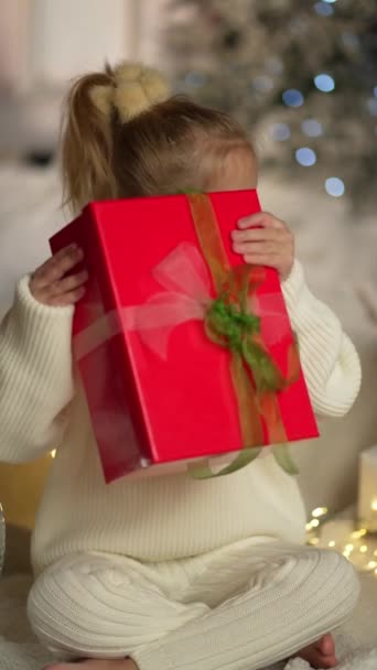 Dětské Vánoce. Dívka třese dárkovou krabicí s lukem a snaží se uhodnout, co je uvnitř. Dítě dostalo novoroční dárek. Veselé Vánoce - Záběry, video