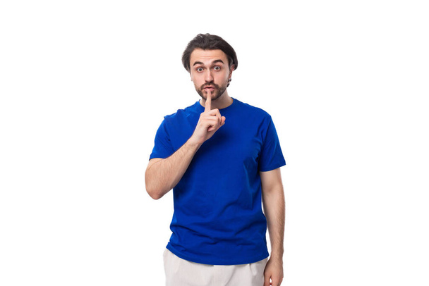 jonge brutale Kaukasische brunette man met een baard in een blauw t-shirt houdt het geheim van business development op een witte achtergrond met kopieerruimte. - Foto, afbeelding