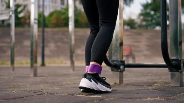 Κοντινό πλάνο των ποδιών του αθλητικού γυμναστή κορίτσι εκτελεί ασκήσεις με μια ελαστική ζώνη για την ενίσχυση των μυών στο πεδίο του αθλητισμού - Πλάνα, βίντεο