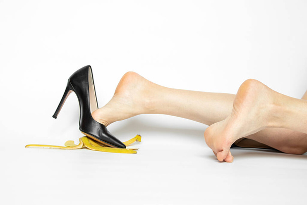 Pés femininos em sapatos de salto alto pretos deslizam em uma casca de banana em um fundo branco, deslizamento e queda, acidente - Foto, Imagem
