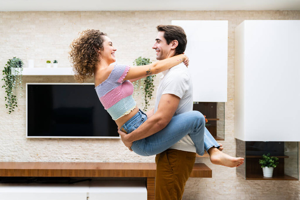 LED TVに対する現代アパートでロマンチックな瞬間を過ごしながら笑顔の若いカップルの側面のビューと幸せにお互いを見る - 写真・画像