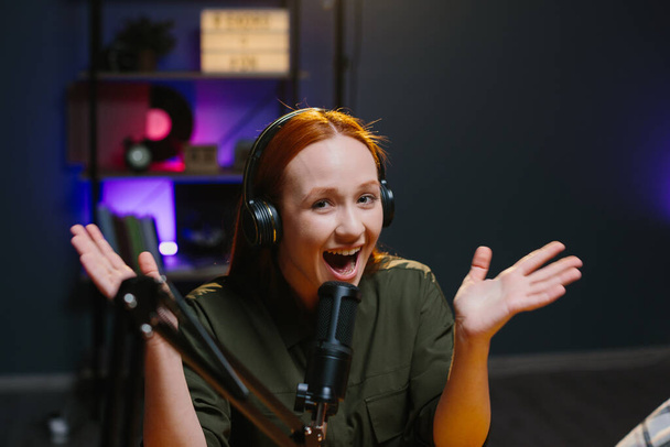 Una chica con auriculares habla expresivamente en un micrófono mientras graba un nuevo episodio de blog. - Foto, Imagen