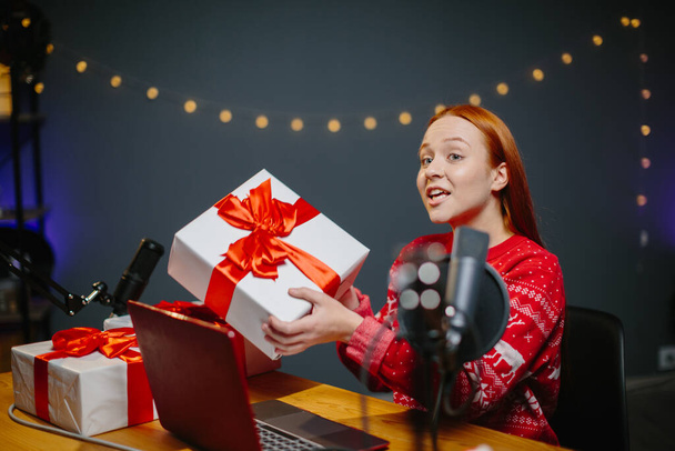 Tytön bloggaaja pitää joululahjaa kanavallaan. Muotokuva tyttö joulun villapaita lahjoja käsissään kotona studio. - Valokuva, kuva
