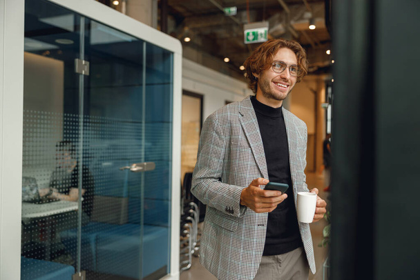 Guapo gerente masculino utilizar el teléfono móvil y beber café, mientras que de pie en el fondo de la oficina - Foto, imagen