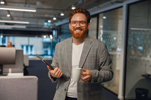 Retrato de belo gerente masculino de pé no fundo do escritório e olha câmera com sorriso - Foto, Imagem