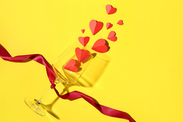 Komposition mit Glas, Schleife und Papierherzen auf gelbem Hintergrund. Valentinstag-Feier - Foto, Bild