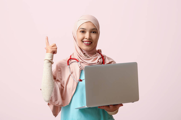 Nainen muslimi lääkäri hijabissa moderni kannettava osoittaa jotain vaaleanpunainen tausta. Maailman hijabipäivän käsite - Valokuva, kuva