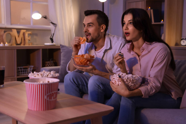 Young couple with potato chips and popcorn watching TV at home in evening - Photo, Image