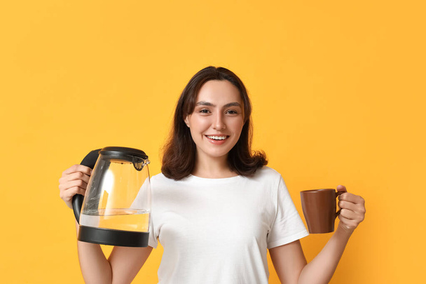 Bella giovane donna con moderno bollitore elettrico e tazza su sfondo giallo - Foto, immagini