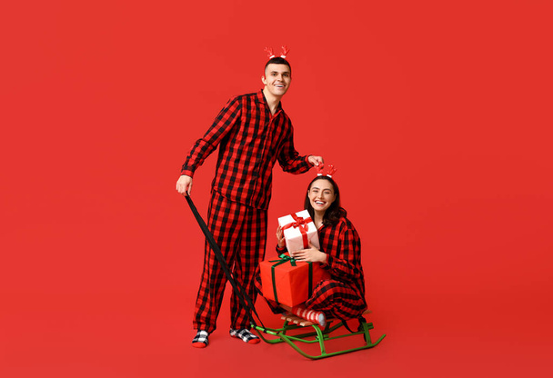 Happy young couple in Christmas pajamas, with sledge and gifts on red background - Φωτογραφία, εικόνα
