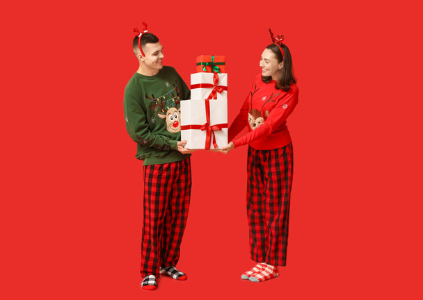 Boldog fiatal pár karácsonyi pizsamában és ajándékokkal a piros háttér - Fotó, kép