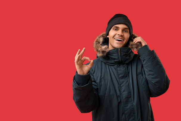 Young man in stylish puffer jacket and hat showing ok gesture on red background - Photo, Image