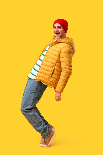Young man in stylish puffer jacket and hat on yellow background - Фото, изображение