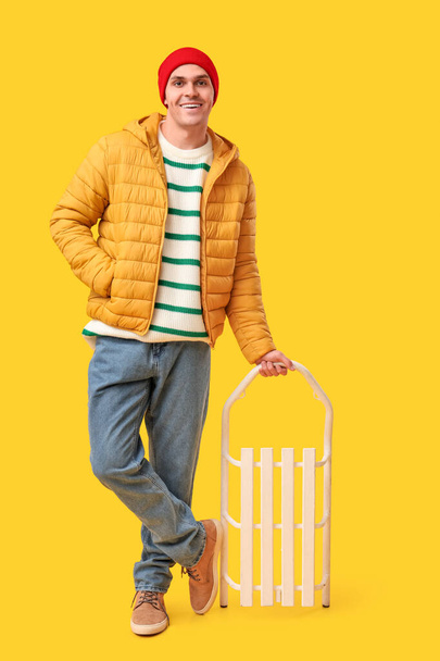 Young man in stylish puffer jacket with sled on yellow background - Foto, Imagem