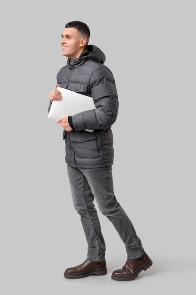 Young man in stylish puffer jacket with modern laptop on grey background - Фото, зображення