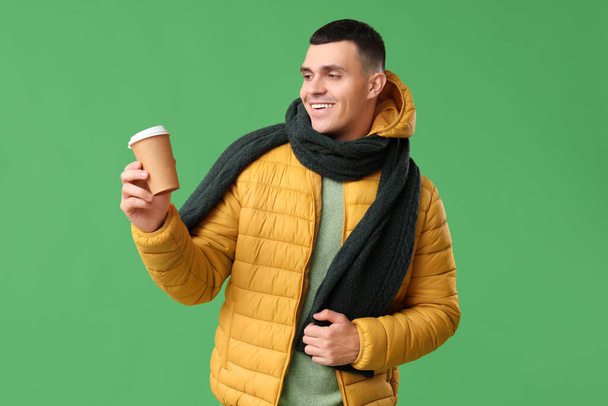 Sarı renkli kabarık ceketli ve atkılı genç adam yeşil arka planda bir fincan kağıt kahve. - Fotoğraf, Görsel