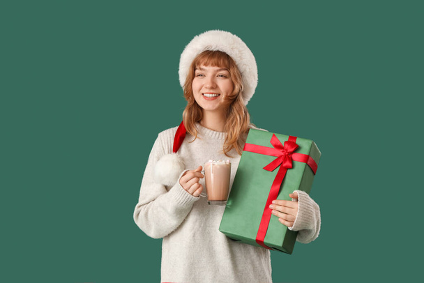 Pretty young woman in Santa hat holding cup of hot chocolate and Christmas gift on green background - Photo, Image