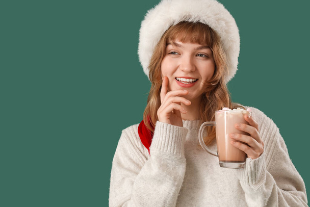 Hübsche junge Frau mit Weihnachtsmütze hält eine Tasse heiße Schokolade mit Marshmallows auf grünem Hintergrund - Foto, Bild