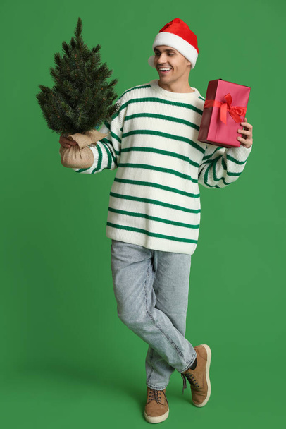 Young man in Santa hat with Christmas tree and gift box on green background - Photo, Image