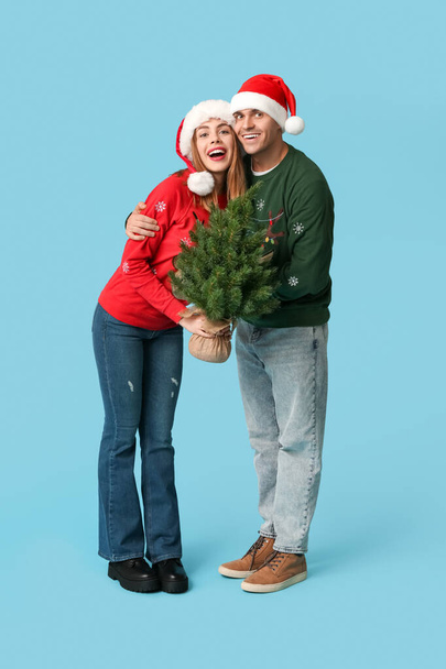 Happy young couple in Santa hats with Christmas tree on blue background - Photo, Image