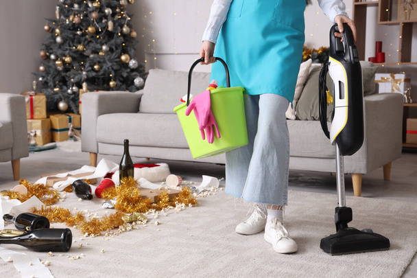 Jovem zelador com suprimentos de limpeza na sala de estar bagunçada após a festa de Ano Novo - Foto, Imagem