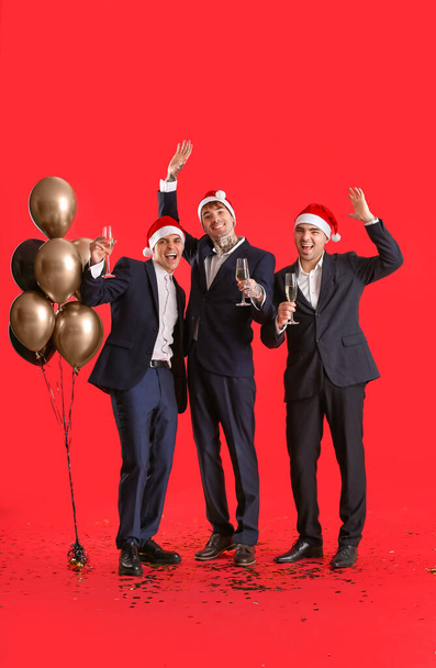 Young men in Santa hats with champagne and balloons on red background. New Year celebration - Photo, Image