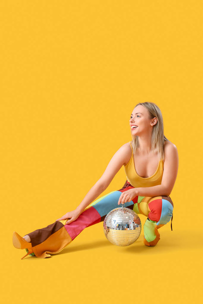 Pretty young woman in retro outfit with disco ball on yellow background - Photo, Image