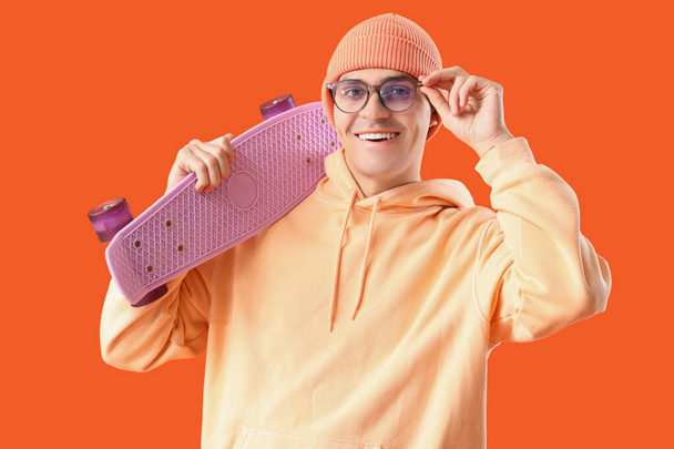 Молодий чоловік в окулярах зі скейтбордом на помаранчевому фоні - Фото, зображення