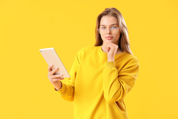 Mujer joven reflexiva en gafas con tableta sobre fondo amarillo - Foto, Imagen